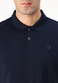 Námornícky polo tričko vyrobené z textúrovanej látky, s klasickým golierom, trojitou gombíkovou légou a jemným logom na ľavej hrudi.