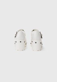 White heeled shoes with a glossy finish, featuring a buckle strap and black studs for detail, set on a thick platform sole.