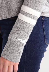 Suéter gris de punto con detalles de franjas blancas en las mangas y un pequeño logo blanco. Combinado con jeans de mezclilla azul oscuro. Textura suave.