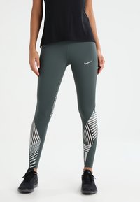 Donna che indossa leggings Nike verde scuro con motivi geometrici bianchi e scarpe da ginnastica nere, in piedi su uno sfondo bianco.