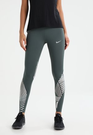 Kobieta ubrana w ciemnozielone legginsy Nike z białymi wzorami geometrycznymi i czarnymi butami sportowymi, stojąca na białym tle.