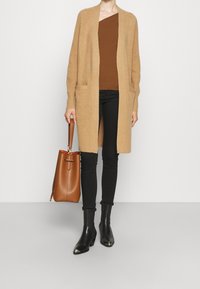 Beige cardigan med öppen framsida och två fickor, lager över en brun topp. Svarta tajta byxor och svarta ankelskor. Brun handväska.