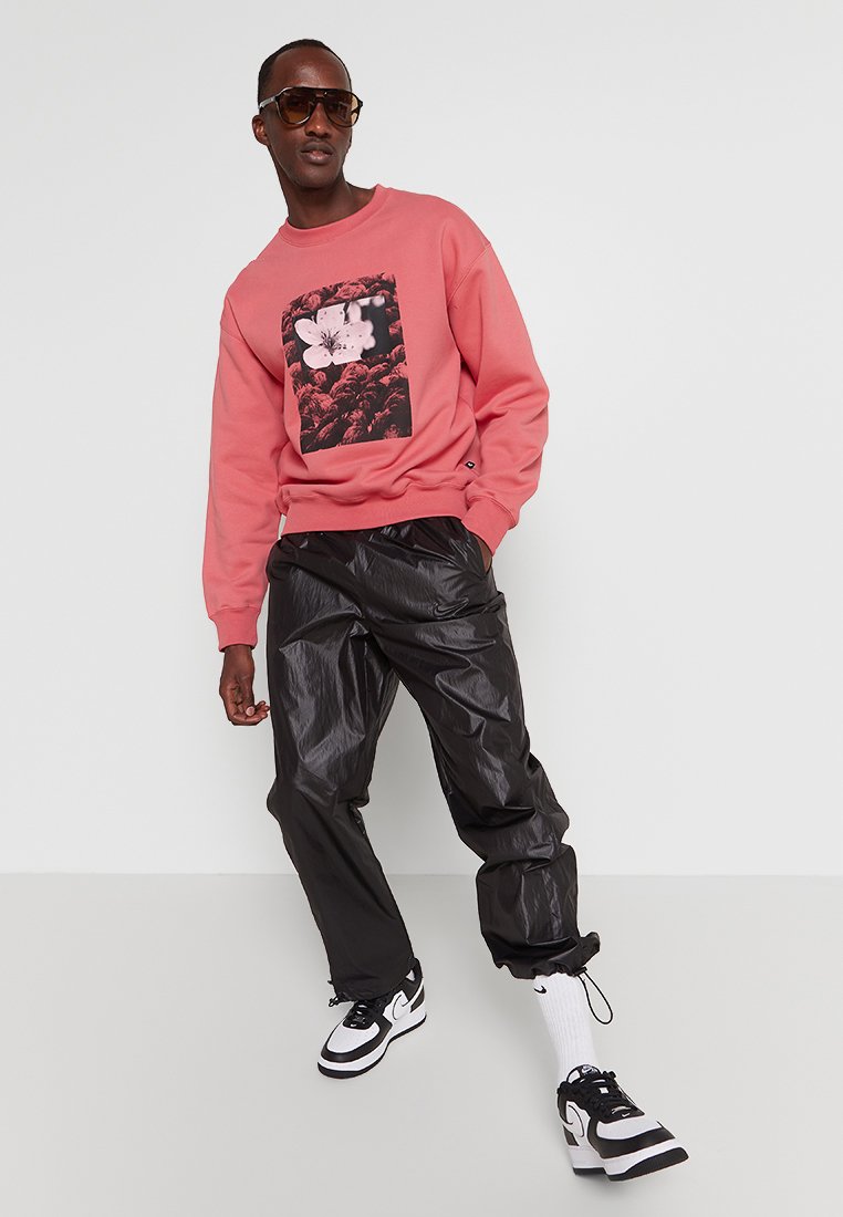 Jasnoróżowa bluza z graficznym wzorem kwiatowym, zestawiona z błyszczącymi czarnymi spodniami cargo i czarno-białymi sneakersami. Na sobie czarne okulary przeciwsłoneczne.
