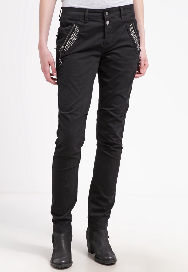 Femme portant un pantalon slim noir avec des poches zippées cloutées et des bottines noires, debout sur un fond blanc.