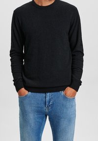 Man som bär en svart långärmad tröja med ribbade manschetter och fåll, samt ljusblå denimjeans, med händerna i fickorna, mot en enfärgad bakgrund.