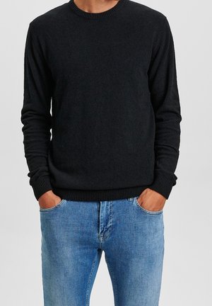 Mann trägt einen schwarzen Pullover mit langen Ärmeln und gerippten Bündchen sowie einen hellblauen Jeans, die Hände in den Taschen, vor einem schlichten Hintergrund.