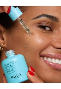 Helles blaues Pipettenfläschchen mit der Aufschrift "KIKO Skin Tech Serum Hydrating Booster", aus dem ein klarer Tropfer Serum auf die Wange eines Models gegeben wird.
