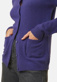 Lila kofta i mjukt stickat material, med knäppning fram, två framfickor och ribbade ärmslut för extra textur.