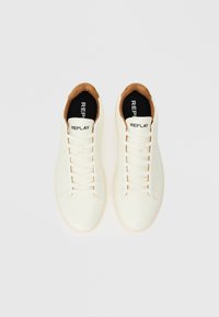 Baskets blanches avec une tige en cuir lisse, des lacets plats et un accent marron au niveau du col. Semelle intérieure noire avec le logo "REPLAY".