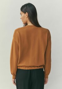 Pulover maro, cu dungi, cu mâneci lungi și croială relaxată, având tiv adunat și textură netedă. Purtat cu pantaloni negri.