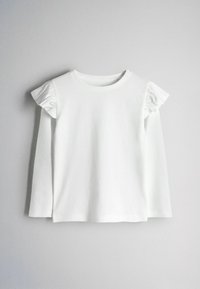 Chemise blanche à manches longues avec des détails à volants sur les épaules, posée à plat sur un fond clair uni.