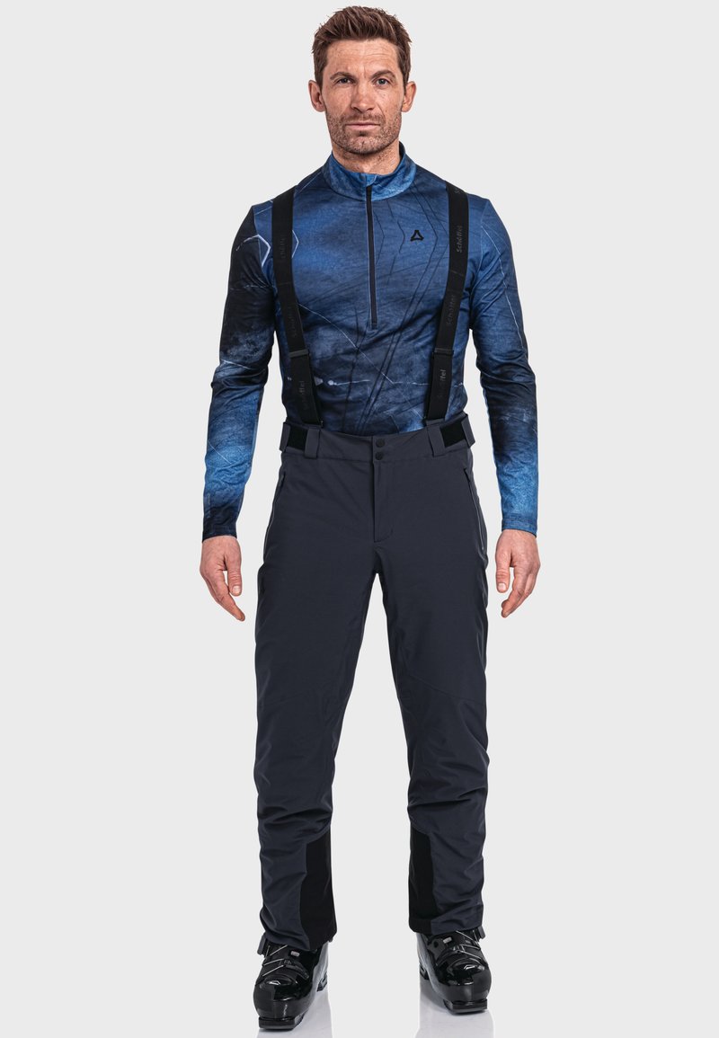 Mann in einem blauen gemusterten Langarmshirt, schwarzen Skihosen mit Trägern und schwarzen Skischuhen steht vor einem einfarbigen Hintergrund.