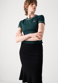 Top ajustat verde cu mâneci scurte, cu margini contrastante și logo brodat, asortat cu o fustă midi neagră cu borduri canalate și tiv evazat.