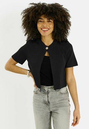 Jeune femme aux cheveux bouclés souriante, portant un cardigan court noir à manches courtes et un jean taille haute gris clair.