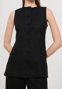 Černá vestička bez rukávů z texturovaného lnu, s vysokým límcem, šesti předními knoflíky a dvěma bočními kapsami. Hladký, minimalistický design.