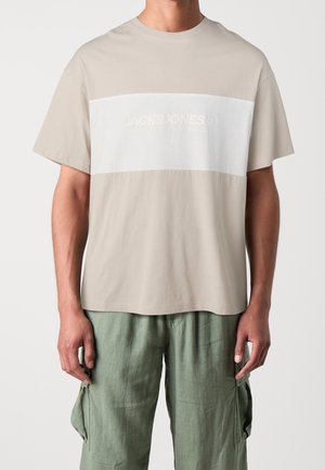 Mężczyzna ubrany w beżowo-biały T-shirt Jack & Jones oraz zielone spodnie bojówki, stojący na jednolitym jasnym tle.