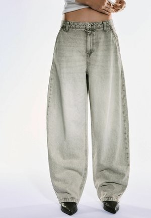 Person wearing wide-leg faded gray jeans and black pointed-toe shoes, hands adjusting waistband, against plain light background.