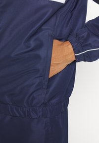 Navy windbreaker with a textured finish, elastic cuffs, and pocket detail. Features white accent stripes for contrast.