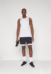 Débardeur de sport blanc avec logo noir, associé à un short noir avec des accents blancs. Le mannequin tient une bouteille d'eau noire et porte des baskets noires.