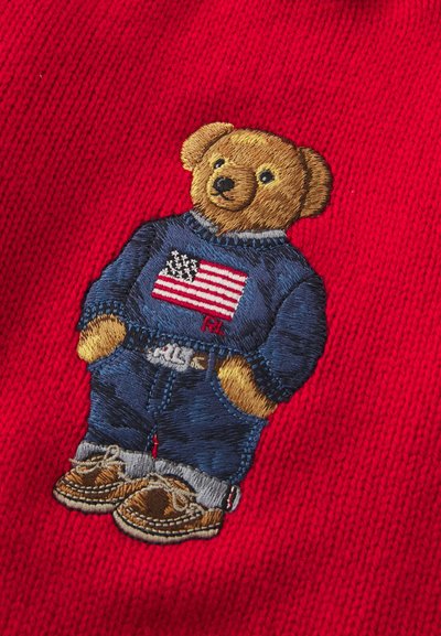 Broderad nallebjörn klädd i en blå tröja med en amerikansk flagga, denimbyxor och bruna skor, mot en röd bakgrund.