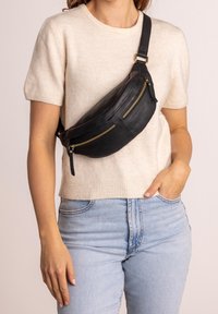 Černá kožená ledvinka s dvěma zipovými kapsami, zakřiveným tvarem, zlatými detaily a nastavitelným popruhem, nošená přes světlý svetr a džíny.