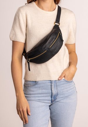 Czarna skórzana nerka z dwiema zapinanymi na zamek kieszeniami, o zaokrąglonym kształcie, ze złotymi akcesoriami i regulowanym paskiem, noszona na jasnym swetrze i dżinsach.