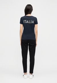 Donkerblauw katoenen T-shirt met een ronde hals en korte mouwen. "ITALIA" in dikke witte letters op de achterkant gedrukt. Zwarte broek en witte sneakers.
