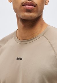 Άνδρας που φοράει μπεζ μπλούζα με στρογγυλή λαιμόκοψη και το λογότυπο "BOSS" τυπωμένο με μαύρα γράμματα στο στήθος, φαίνεται από το πιγούνι μέχρι το πάνω μέρος του στήθους.