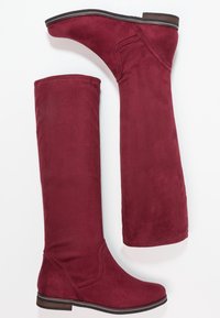 Bottes bordeaux montant jusqu'au genou en suède doux, avec un bout légèrement pointu et un talon bas empilé. Détail de coutures le long de la tige.