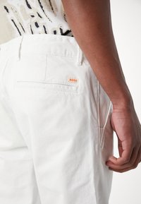 White cotton pants with a back pocket, featuring a small orange "BOSS" label. Smooth texture, classic fit, and minimal design details.