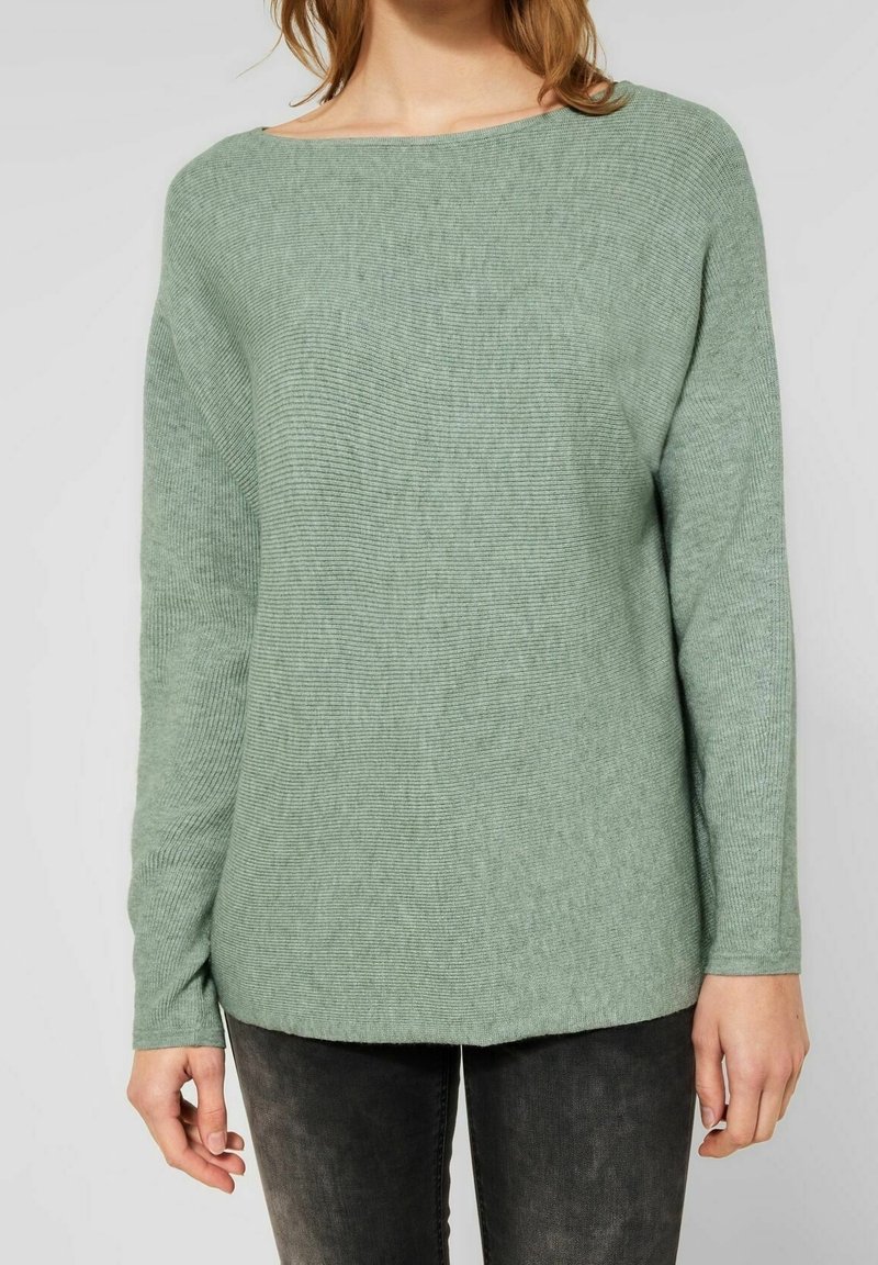 Femme portant un pull à côtes vert clair à manches longues et un pantalon gris foncé, debout devant un fond clair uni.