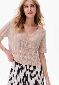 Beżowy sweter włóczkowy z dekoltem w kształcie V, krótkimi rękawami z mankietami i detalami w postaci guzików. Wzór zawiera otwartą robótkę w geometrycznym designie.