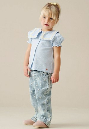 Unge barn med blondt hår iført en blåstribet top med stor krave og lyseblå jeans med tusindfuglemønstre samt lyserøde hjemmesko.