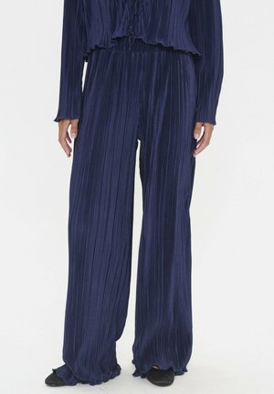 Marineblaue, weit geschnittene Hose mit vertikalen Falten, hohe Taille und strukturiertem Stoff. Komplettiert mit schwarzen Slippers.