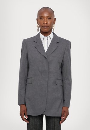 Sivý, prispôsobený blejzer s vykrojeným klopom, dvoma prednými vreckami a jedným vreckom na hrudi; nosený cez bielu košeľu s vzorovaným golierom.