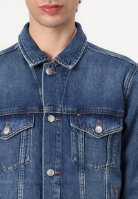 Denimjakke i middels blå nyanse med klassisk krage, to brystlommer, sølvfargede metallknapper og falmede teksturdeler.