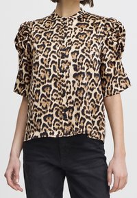 Femme portant un haut à manches courtes en imprimé léopard avec des boutons noirs et un pantalon noir, sur un fond uni.