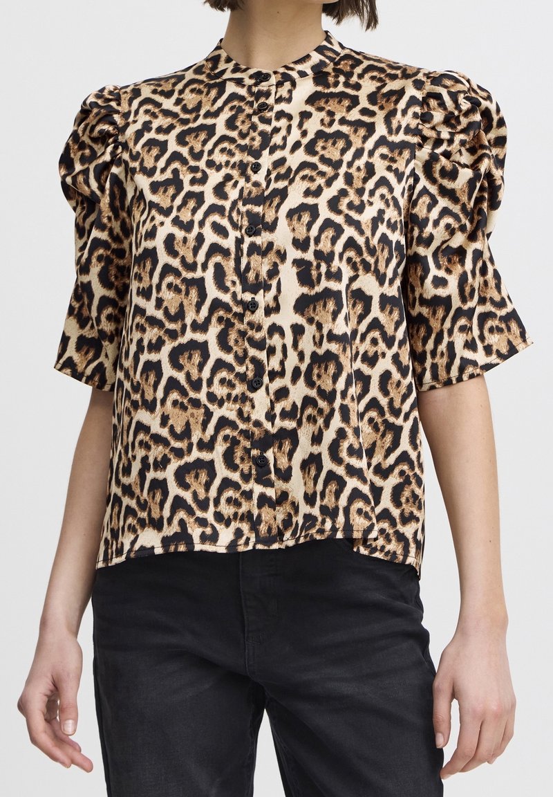 Femme portant un haut à manches courtes en imprimé léopard avec des boutons noirs et un pantalon noir, sur un fond uni.