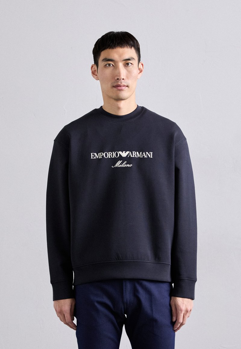 Sweatshirt bleu marine avec col ras du cou, présentant le texte blanc "Emporio Armani Milano" sur le devant et des poignets et un ourlet côtelés.