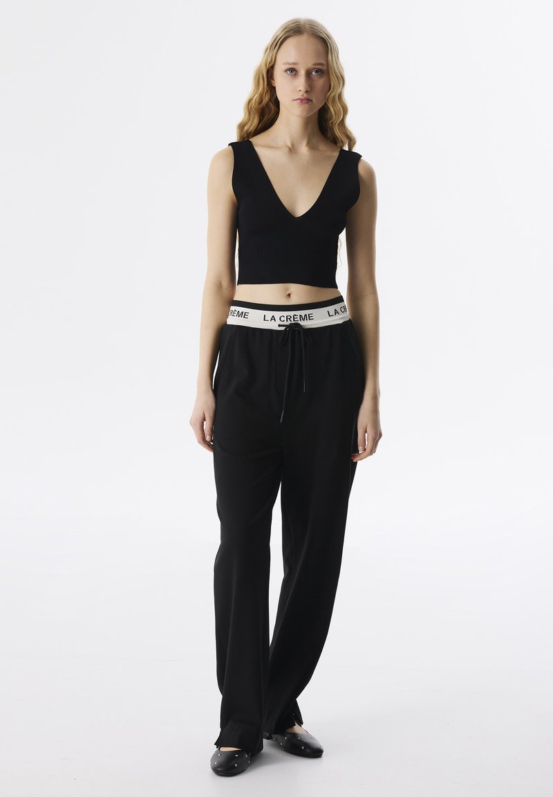 Schwarzes geripptes Crop-Top mit V-Ausschnitt, kombiniert mit lockeren schwarzen Hosen mit einem weißen elastischen Bund mit Markenlogo. Das Modell steht vor einem weißen Hintergrund.
