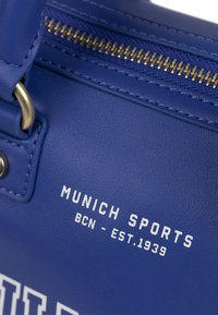 Plava kožna torba s zlatnim patentnim zatvaračem, bijelim otisnutim tekstom "MUNICH SPORTS BCN - EST.1939" sprijeda i glatkom teksturom.