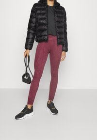 Svart pufferjacka, åtsittande svart t-shirt, vinröda mönstrade leggings och svarta sportskor, med en liten svart väska och en design med avsmalnande ben.