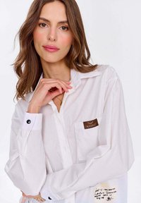Biała koszula na guziki z długimi rękawami, z kieszenią na piersi z brązową metką, czarnymi akcentami guzika oraz odręcznymi detalami na mankietach.