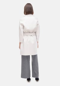Lungo cappotto crema con vita cinturata, vestibilità su misura e maniche 3/4; abbinato a pantaloni grigi larghi e scarpe nere a punta.