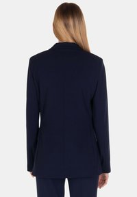 Blazer blu navy con vestibilità sartoriale, rever a punta, maniche lunghe e texture liscia. Presenta dettagli di cucitura sul retro e un design minimalista.