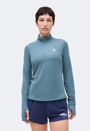Person iført en blå quarter-zip langærmet top og marineblå shorts med teksten "LONDON", stående mod en ensartet lys baggrund.