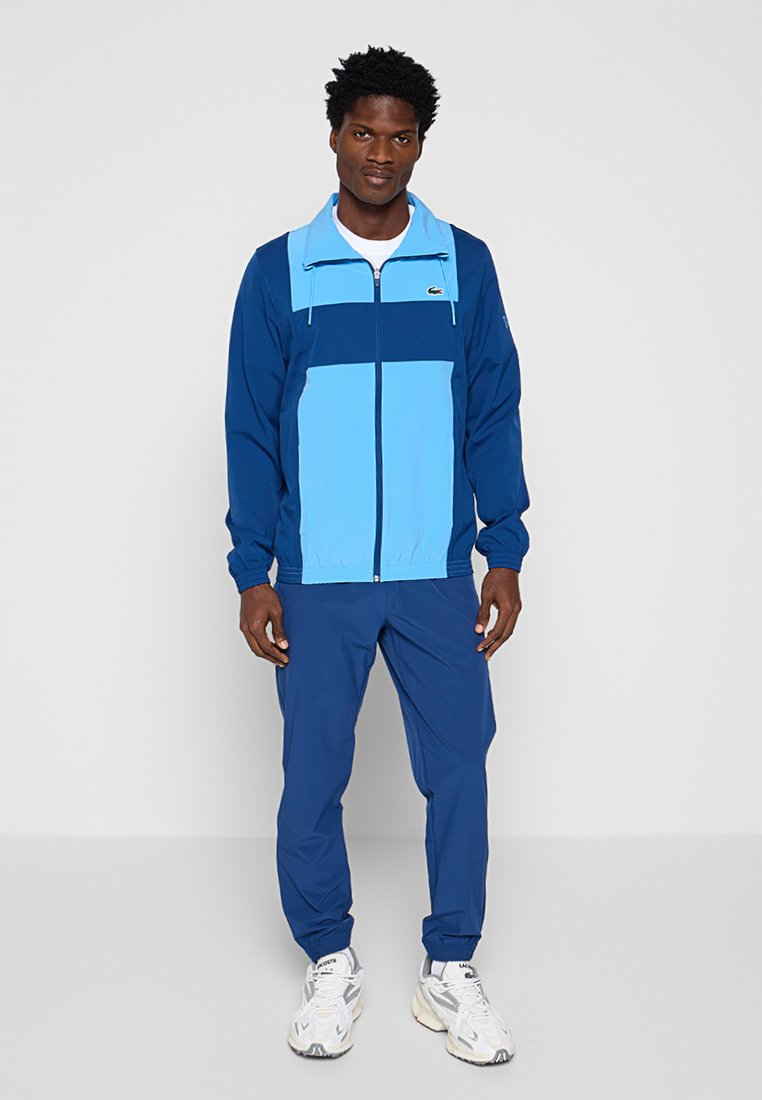 Uomo che indossa una giacca a zip blu a blocchi di colore abbinata a pantaloni blu e sneaker bianche sportive, in piedi su uno sfondo semplice.