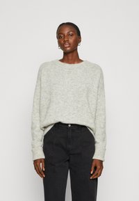 Šviesiai pilkas megztinis su laisvu kirpimu, apvaliu iškirpimu ir raukšlėmis prie rankogalių, derinamas su tamsiomis aukštai užkeliamomis kelnėmis.