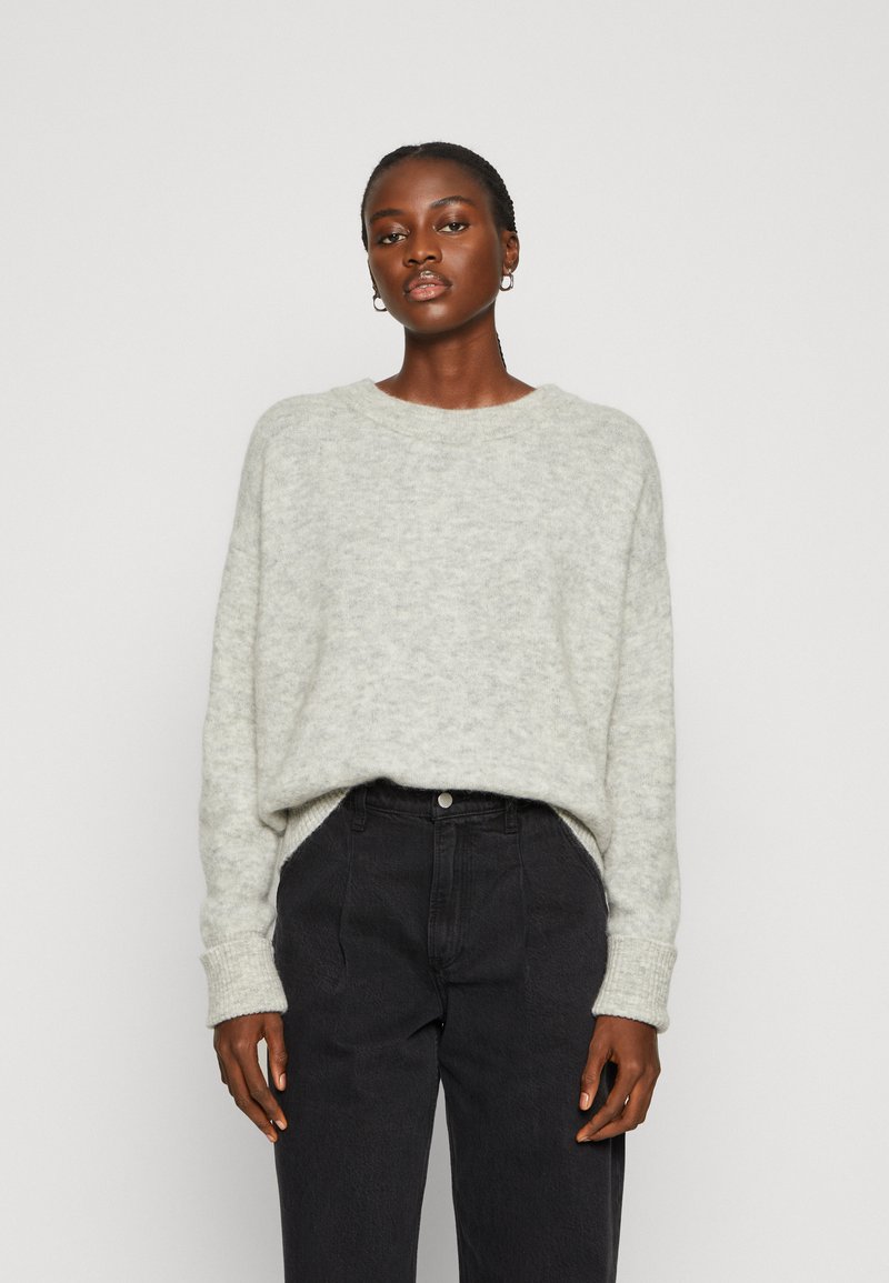 Šviesiai pilkas megztinis su laisvu kirpimu, apvaliu iškirpimu ir raukšlėmis prie rankogalių, derinamas su tamsiomis aukštai užkeliamomis kelnėmis.