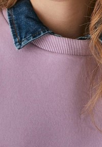 Nära bild av en person som bär en lavendelfärgad ribbad tröja under en blå denimskjortkrage, med långt ljusbrunt hår delvis synligt.